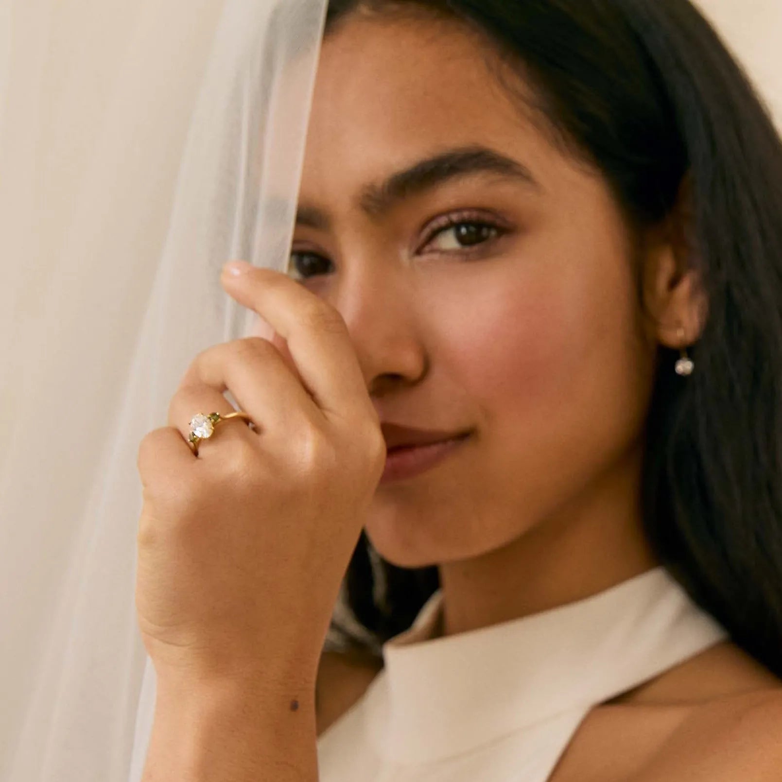 Woman wearing a white dress with a veil and a diamond engagement ring with green touramlines, posing against a plain background