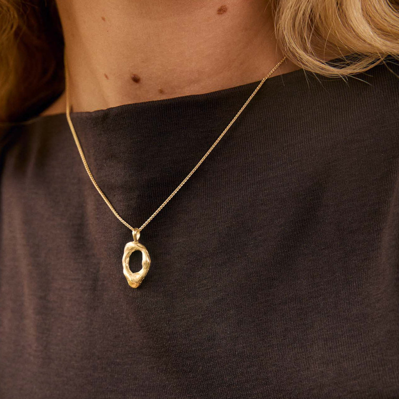 A close-up lifestyle shot of a woman wearing a delicate gold necklace with an abstract, textured oval pendant. She is wearing a dark brown sleeveless top, and her blonde hair is visible against a warm, softly lit background.