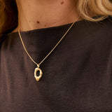A close-up lifestyle shot of a woman wearing a delicate gold necklace with an abstract, textured oval pendant. She is wearing a dark brown sleeveless top, and her blonde hair is visible against a warm, softly lit background.