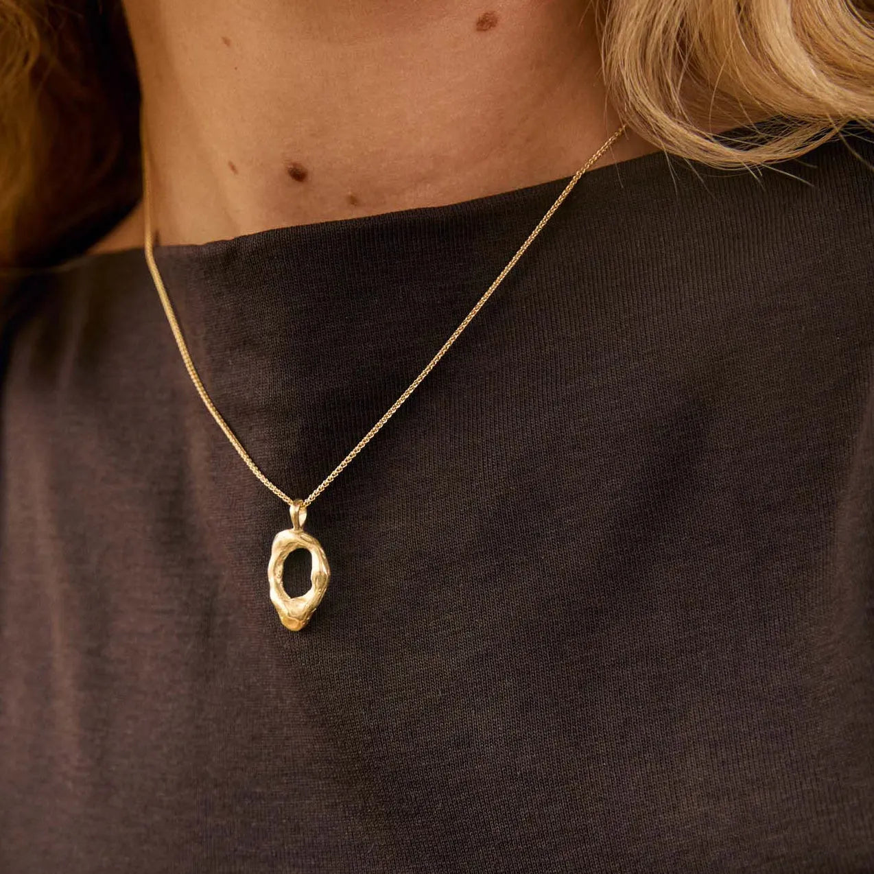 A close-up lifestyle shot of a woman wearing a delicate gold necklace with an abstract, textured oval pendant. She is wearing a dark brown sleeveless top, and her blonde hair is visible against a warm, softly lit background.