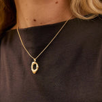 A close-up lifestyle shot of a woman wearing a delicate gold necklace with an abstract, textured oval pendant. She is wearing a dark brown sleeveless top, and her blonde hair is visible against a warm, softly lit background.