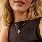 A close-up lifestyle shot of a woman wearing a delicate silver pendant necklace with a textured, openwork oval design. She is wearing a dark brown sleeveless top, and her blonde hair is visible against a warm, neutral background.