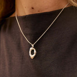 Close up of a silver necklace with molten open oval pendant worn on a womans neck against a brown cotton high-neck top