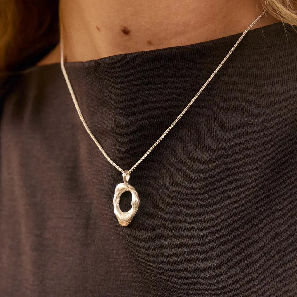 Close up of a silver necklace with molten open oval pendant worn on a womans neck against a brown cotton high-neck top