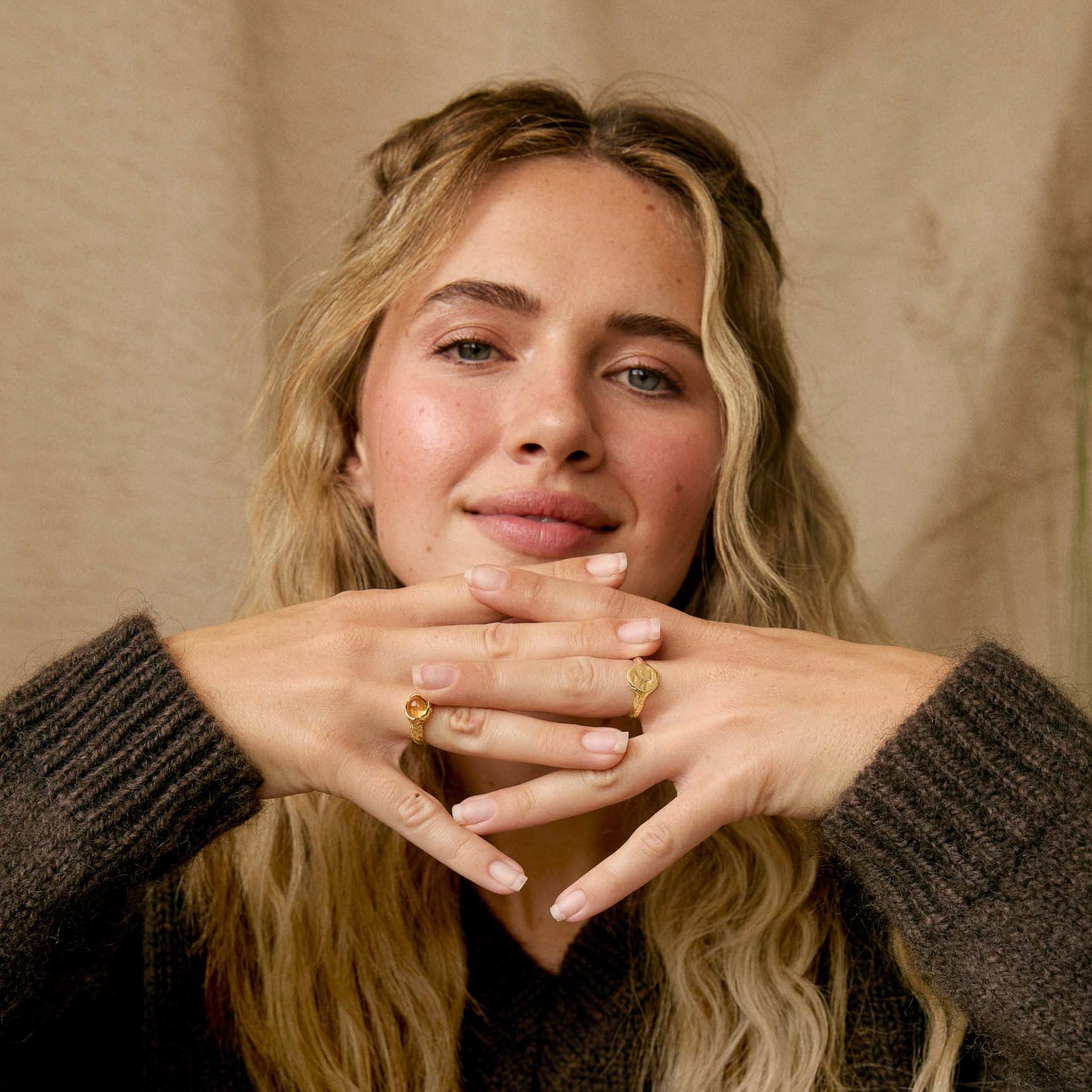 A woman with blonde hair wearing a dark brown ribbed knit sweater and a white ruffled skirt, sitting cross-legged and posing with her hands together to display gold rings.