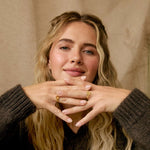 A woman with blonde hair wearing a dark brown ribbed knit sweater and a white ruffled skirt, sitting cross-legged and posing with her hands together to display gold rings.