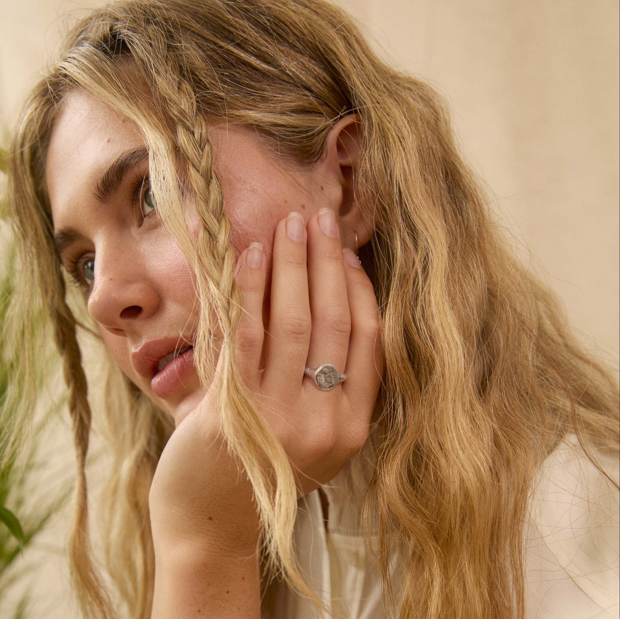 A close-up lifestyle shot of a woman with blonde hair wearing a silver signet ring on her finger, set against a soft beige background.