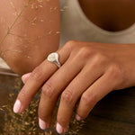 A close-up lifestyle shot of a woman wearing a silver ring with a oval-shaped signet ring on her finger. She is leaning on a dark wooden railing with dried grass in the foreground.
