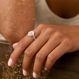 A close-up lifestyle shot of a woman wearing a silver ring with a oval-shaped signet ring on her finger. She is leaning on a dark wooden railing with dried grass in the foreground.