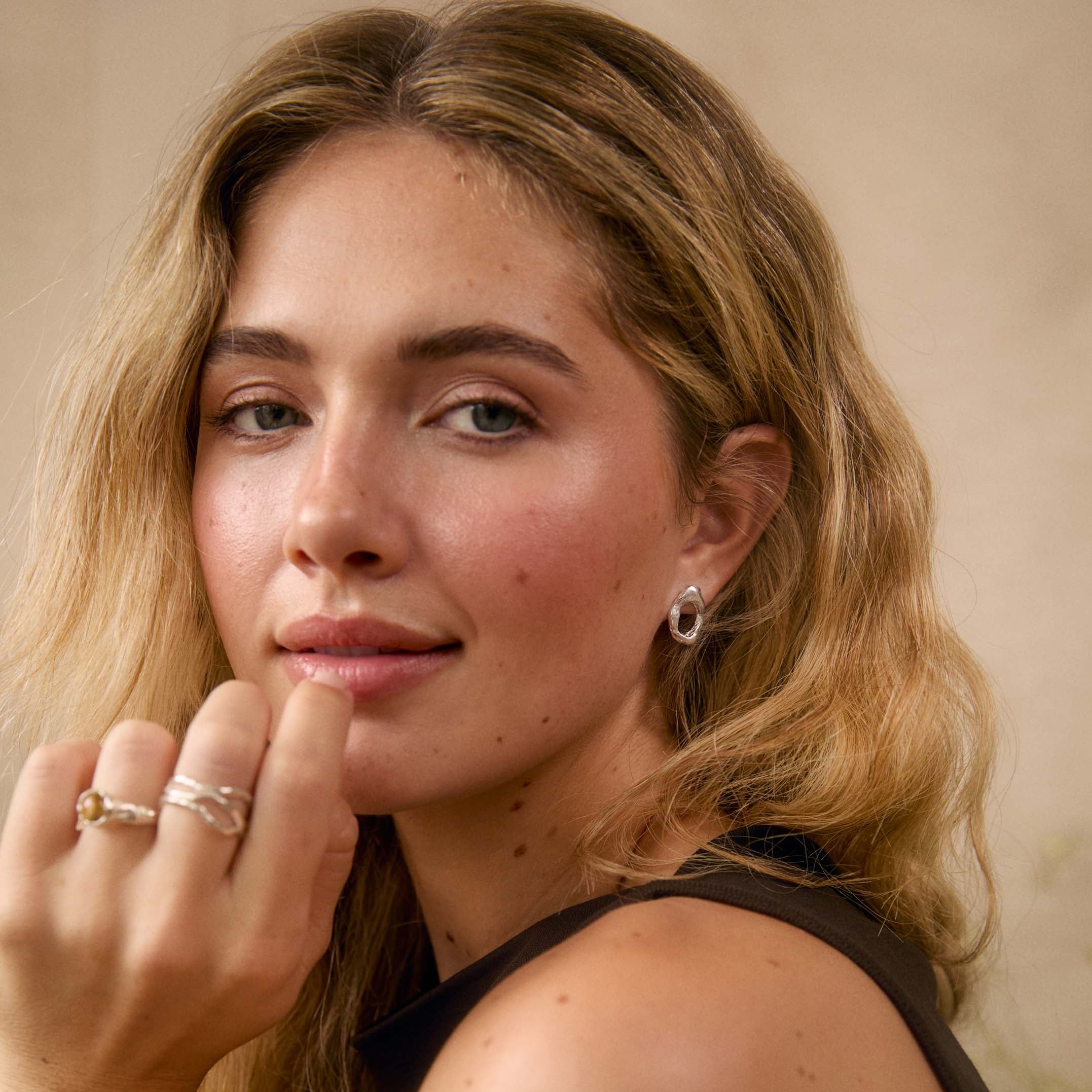 A close-up portrait of a woman with blonde hair wearing silver rings on her fingers and a silver hoop earring, wearing a black sleeveless top against a warm beige background.
