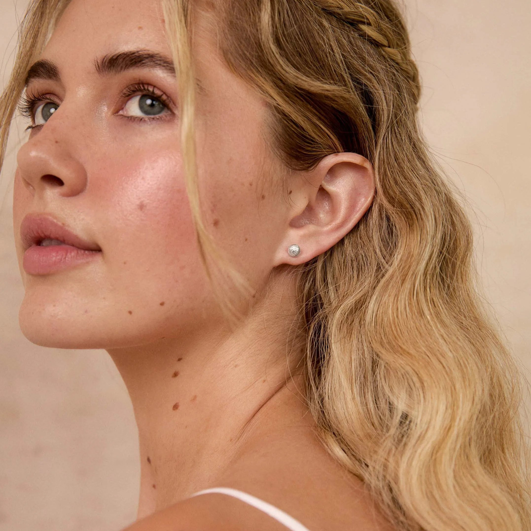 A close-up side profile of a blonde woman with wavy hair, wearing a delicate silver stud earring, against a warm beige background.
