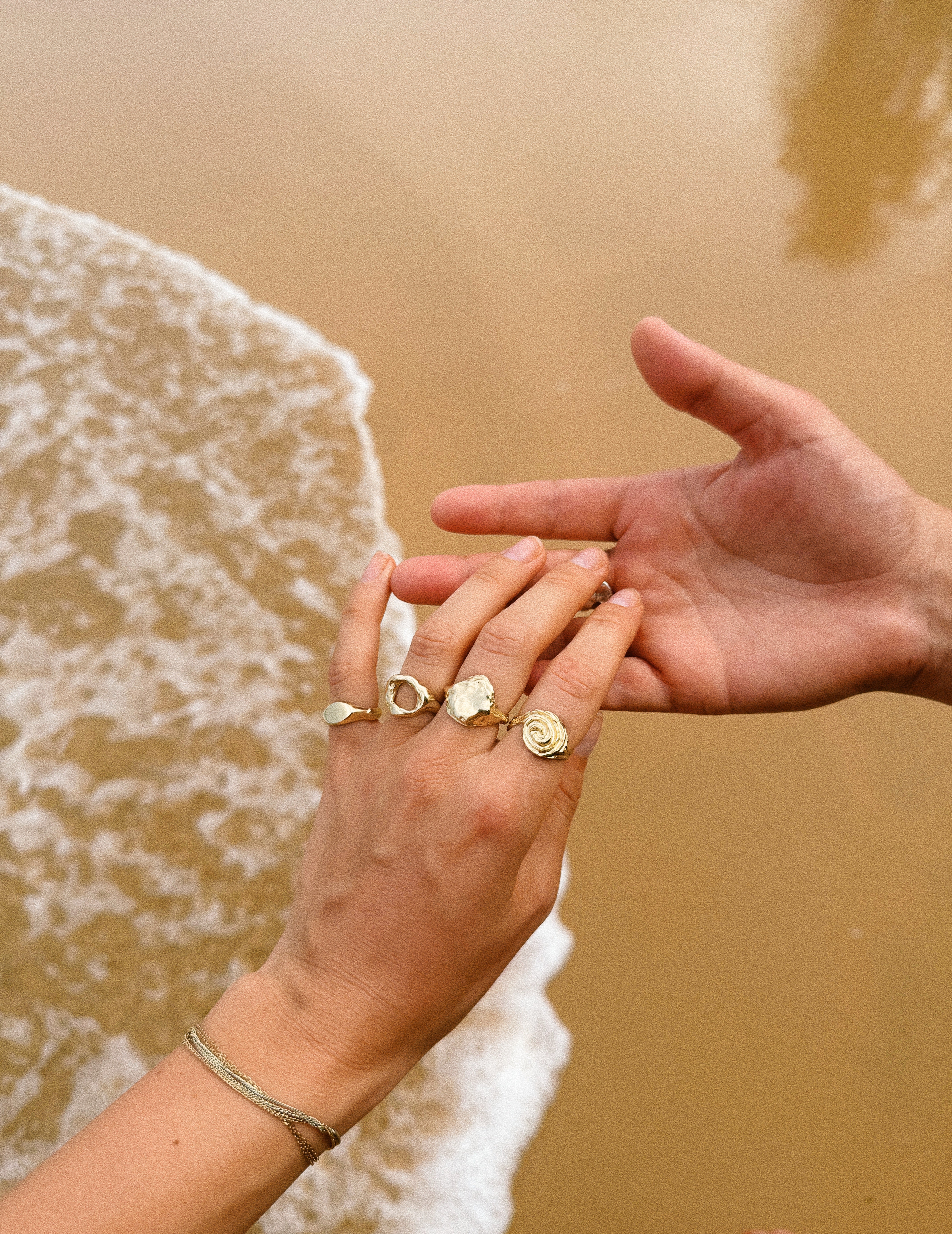 Two womens hands wearing rings made by LGBT owned jewellery brand held together on a sandy beach.