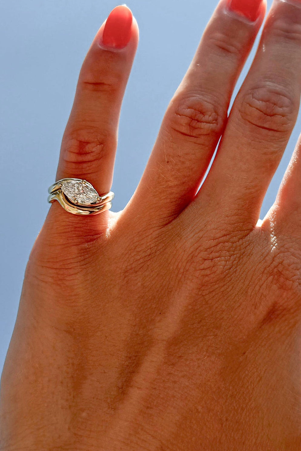 A close-up of a hand with coral-pink nail polish wearing a silver ring with a clear gemstone, set against a bright blue sky.
