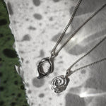 Two silver pendant necklaces, one with an abstract organic ring pendant and the other with a rose-shaped pendant, displayed on a textured white paper surface with leaf shadows.
