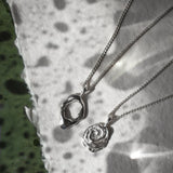 Two silver pendant necklaces, one with an abstract organic ring pendant and the other with a rose-shaped pendant, displayed on a textured white paper surface with leaf shadows.