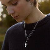Close-up of a woman wearing a silver acorn and oak leaf necklaces and earrings with a blurred natural background