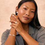 A woman with dark hair wearing a gray button-up shirt models silver jewelry, including a silver sun earring, a silver signet ring, and a silver cuff bracelet, against a textured beige background.
