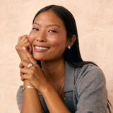 A smiling woman with long dark hair poses with her hands clasped, wearing a gray button-up shirt and delicate jewelry, set against a textured beige wall.