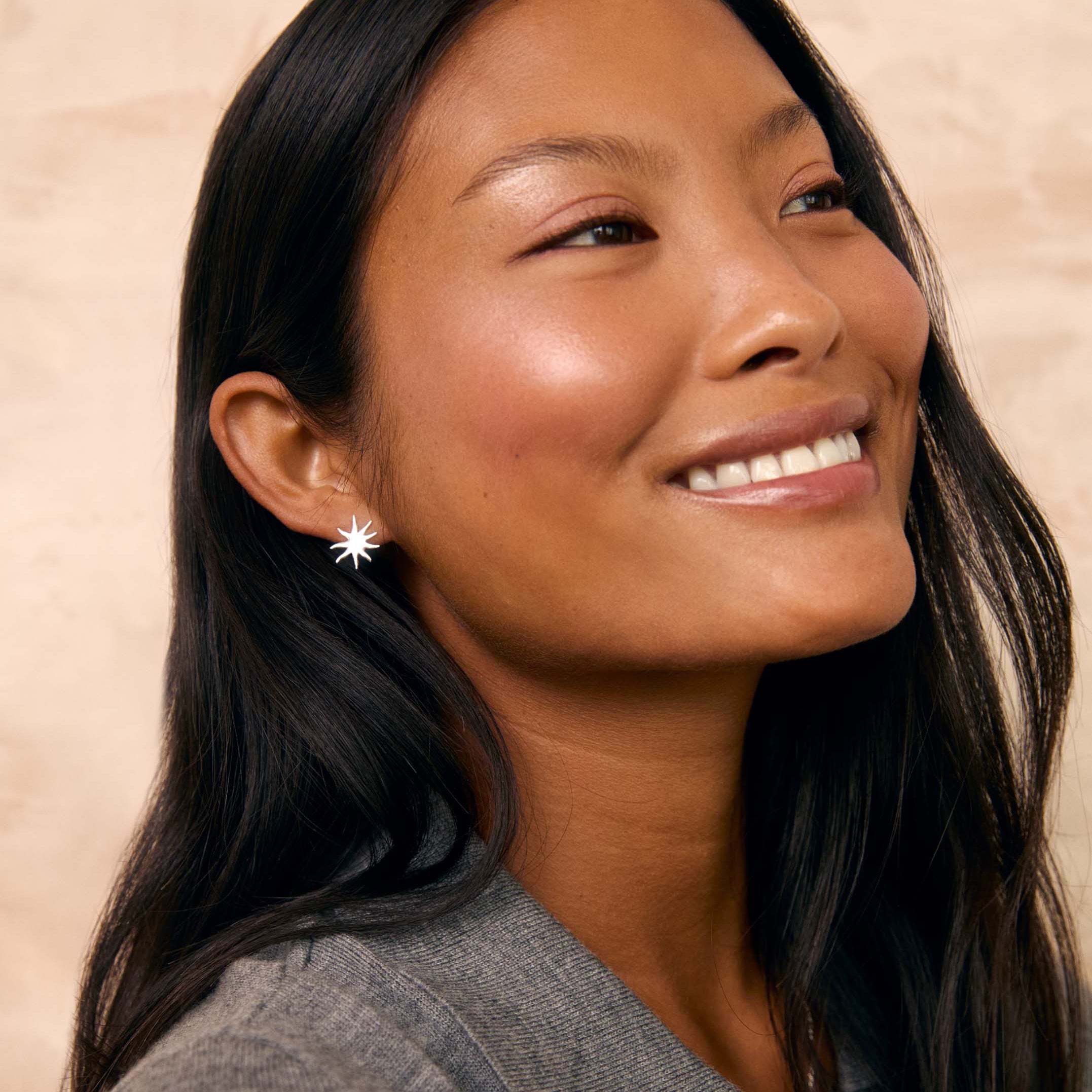A smiling woman with long dark hair wearing a small silver starburst stud earring, photographed against a warm beige textured background.