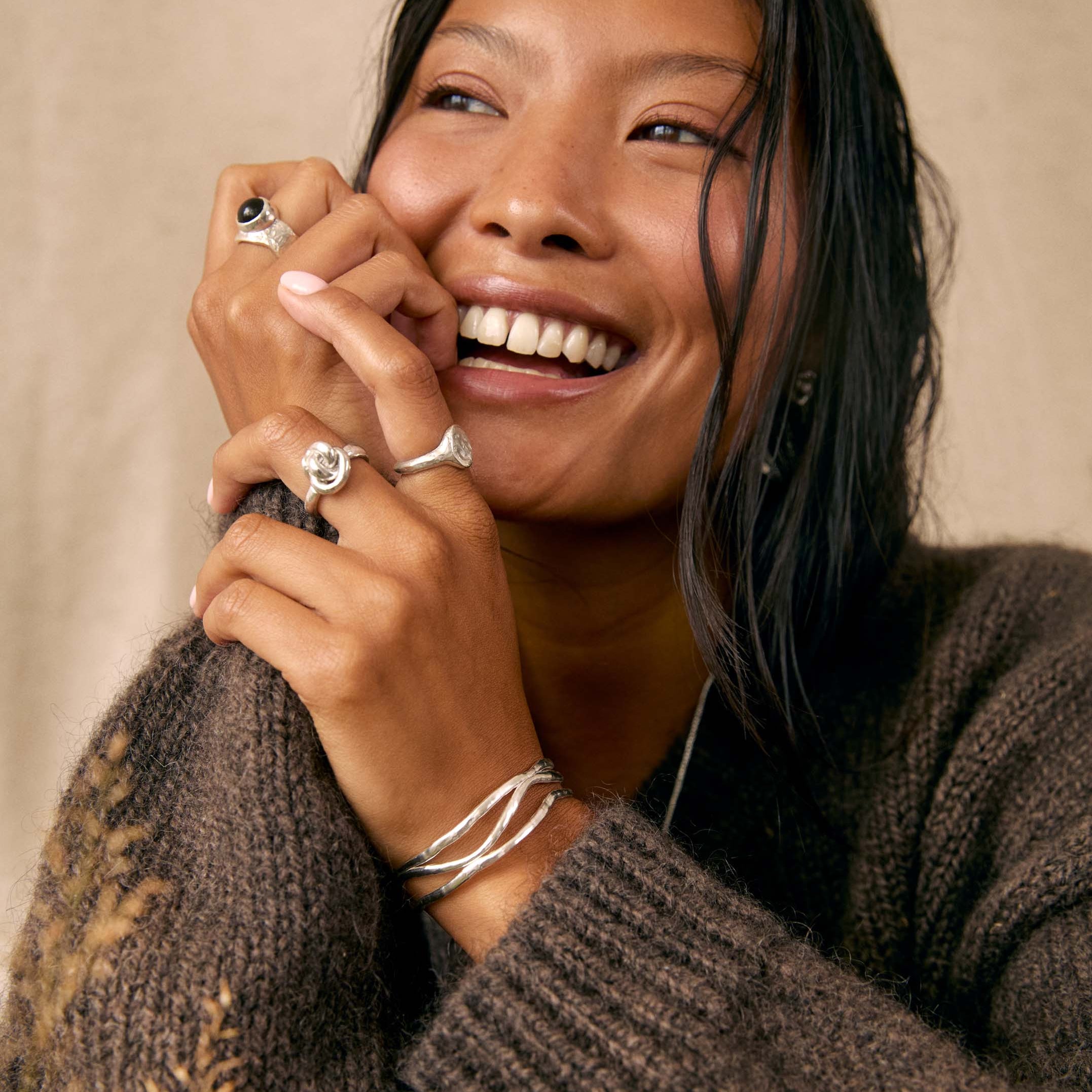 A smiling woman with dark hair wears multiple silver rings and a silver wrap bracelet, posing in a cozy brown knit sweater against a neutral beige background.