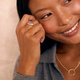 A close-up lifestyle shot of a woman wearing a silver signet ring on her finger and a delicate silver circle necklace, smiling against a soft beige background.