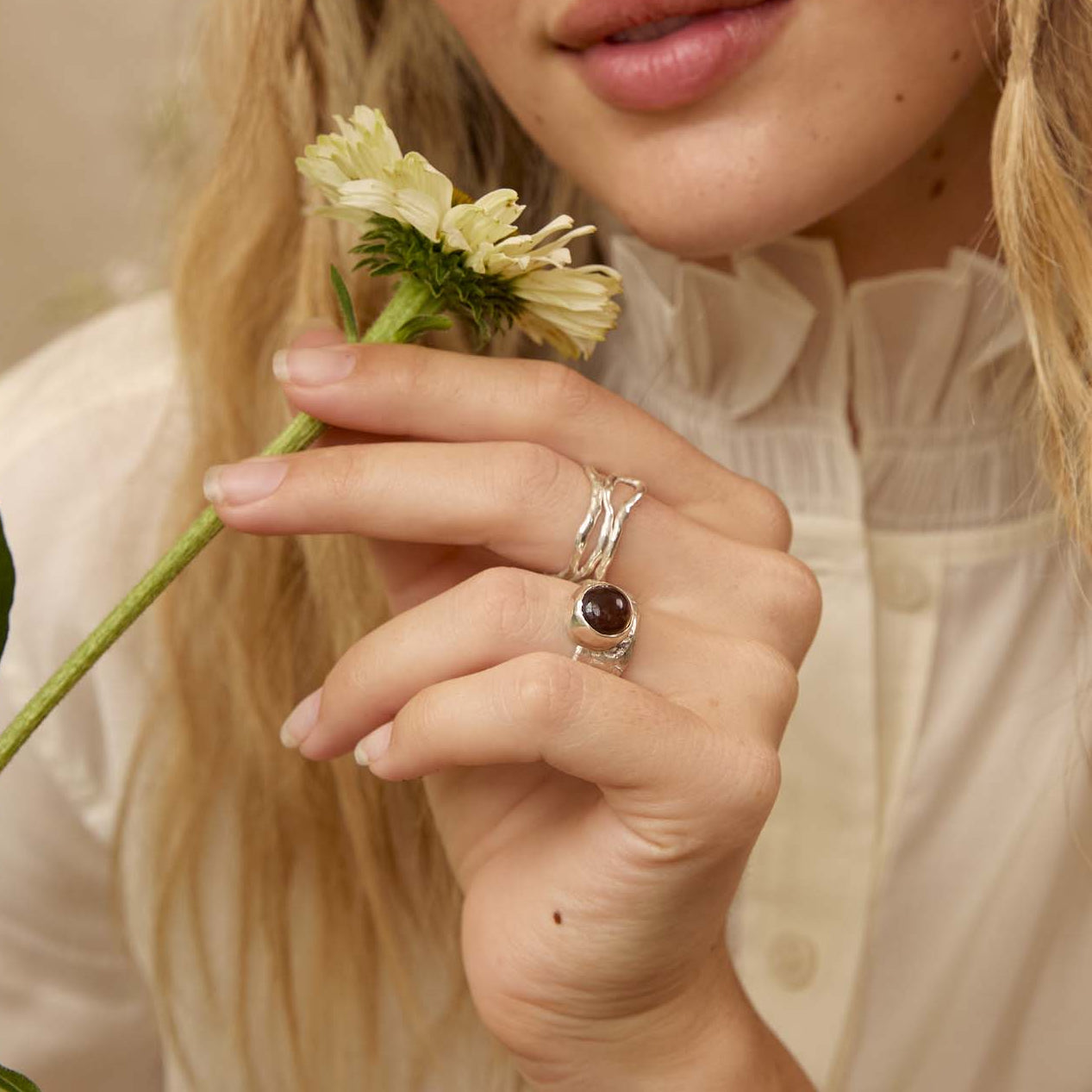 A woman with long wavy blonde hair wearing a cream-colored blouse and a silver ring with a dark oval stone, holding a small white flower. The background is a soft beige wall with dried floral elements.