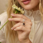 A woman with long wavy blonde hair wearing a cream-colored blouse and a silver ring with a dark oval stone, holding a small white flower. The background is a soft beige wall with dried floral elements.