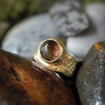 A handmade gold ring with a round, faceted brown gemstone, nestled among natural wet rocks.