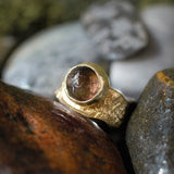 A handmade gold ring with a round, faceted brown gemstone, nestled among natural wet rocks.