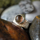 A handmade silver ring with a round, faceted brown gemstone, nestled among natural wet rocks.