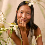 A woman with long dark hair sits among white flowers, wearing a sleeveless light top and striped shorts, with a silver ring on one finger. The background is a soft beige fabric.