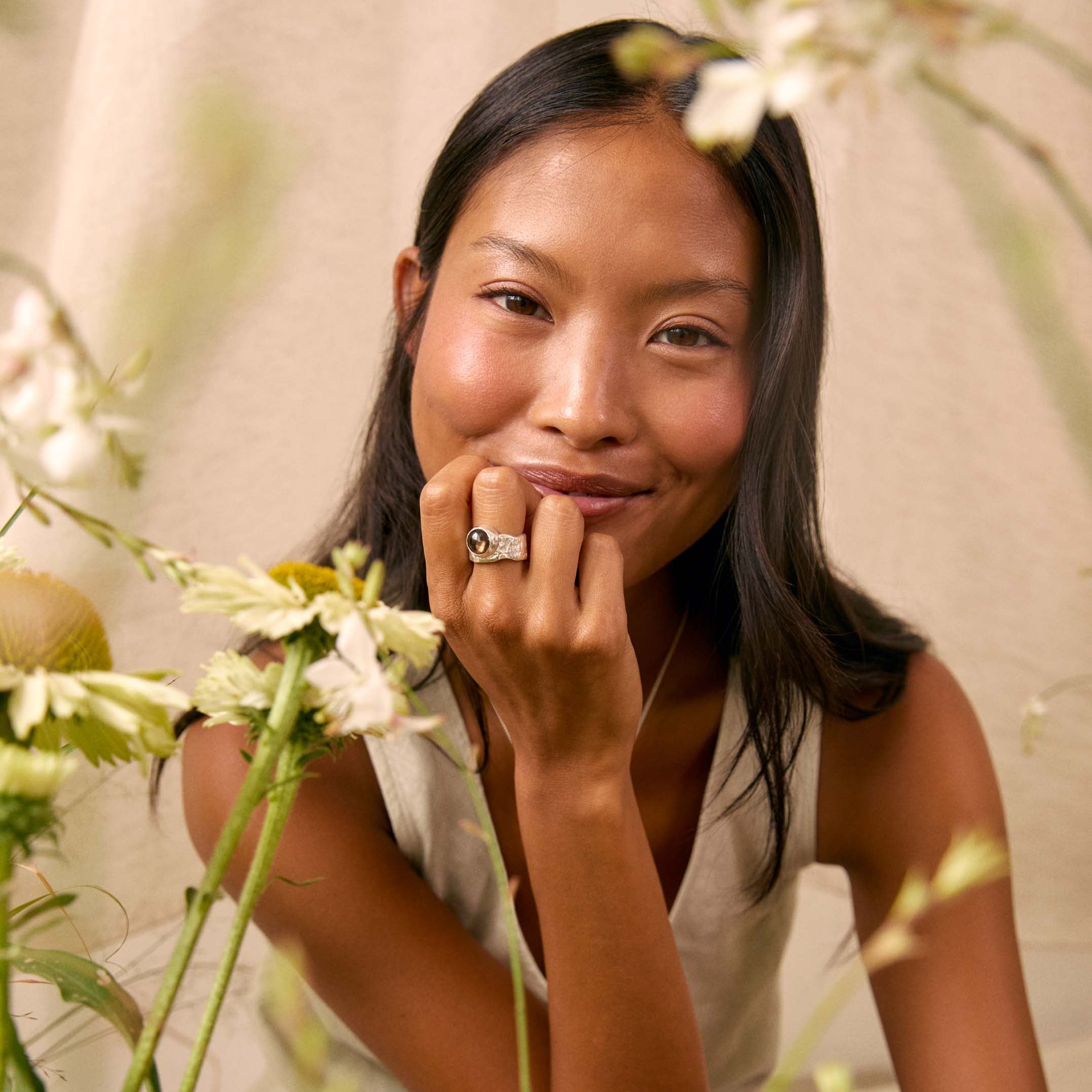 A woman with long dark hair sits among white flowers, wearing a sleeveless light top and striped shorts, with a silver ring on one finger. The background is a soft beige fabric.