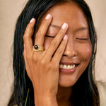 A close-up lifestyle shot of a woman with dark hair smiling and covering part of her face with her hand, wearing a gold ring with a dark round stone.