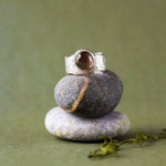 A silver ring with a round brown gemstone is displayed on a stack of three smooth stones against a soft olive green background.