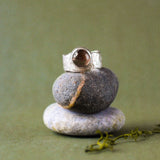 A silver ring with a round brown gemstone is displayed on a stack of three smooth stones against a soft olive green background.
