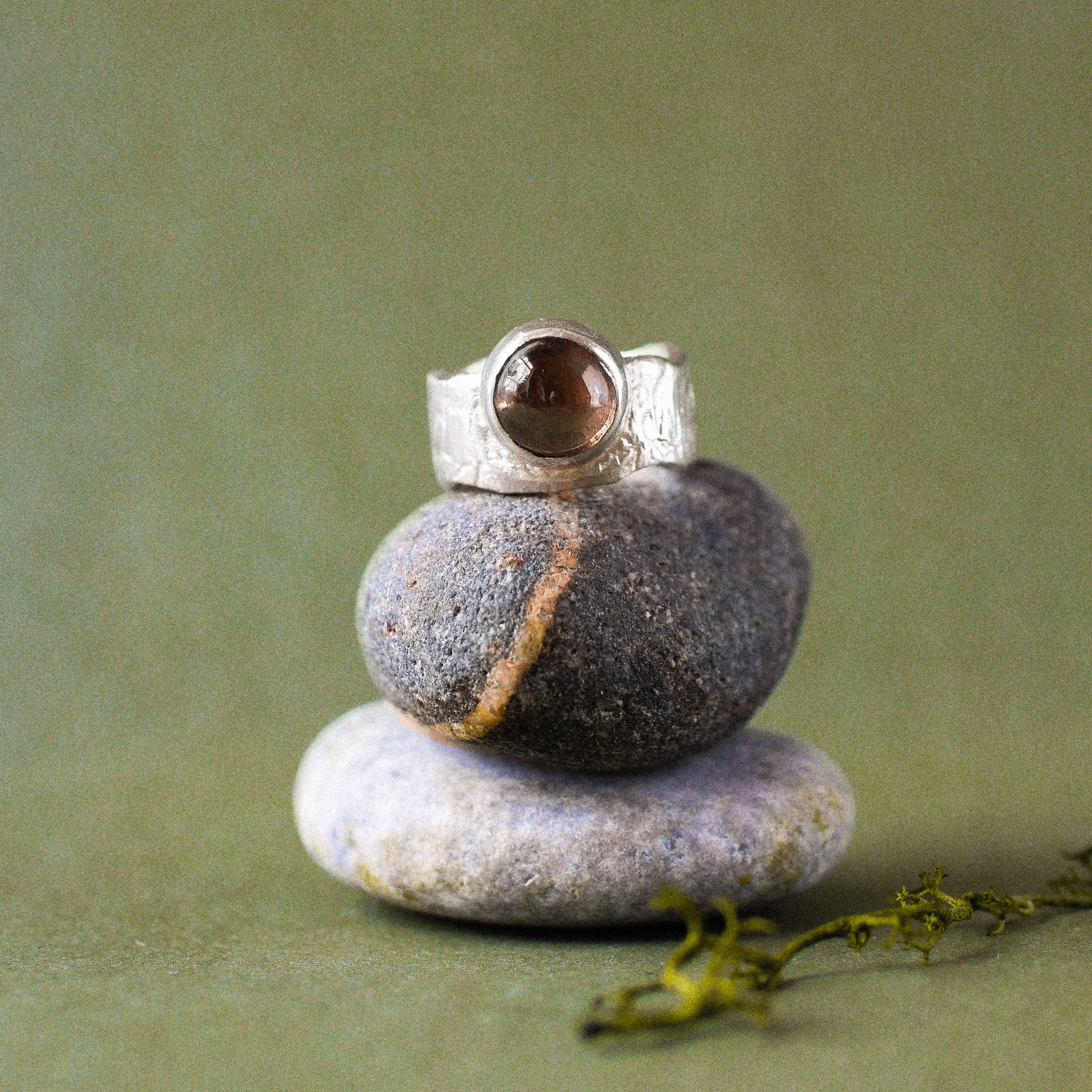 A silver ring with a round brown gemstone is displayed on a stack of three smooth stones against a soft olive green background.