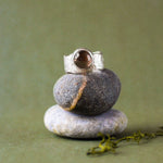 A silver ring with a round brown gemstone is displayed on a stack of three smooth stones against a soft olive green background.