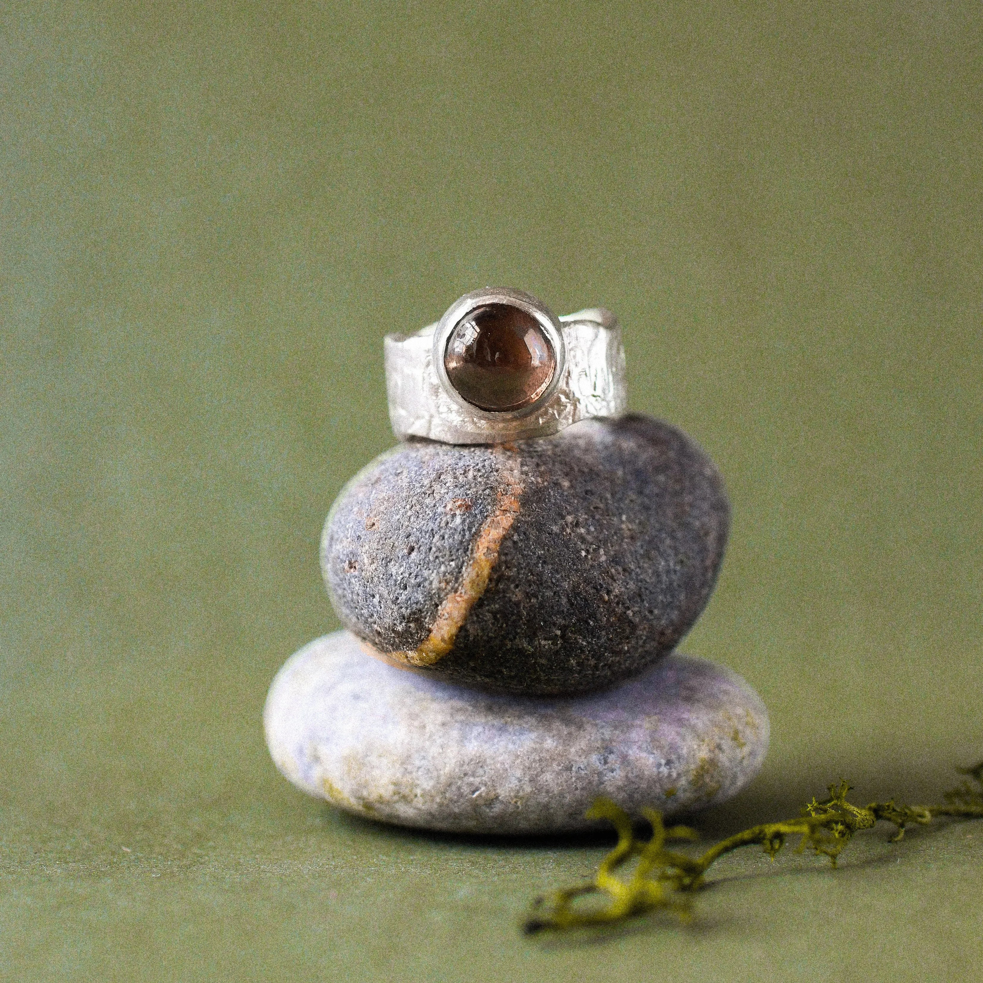 A silver ring with a round brown gemstone is displayed on a stack of three smooth stones against a soft olive green background.