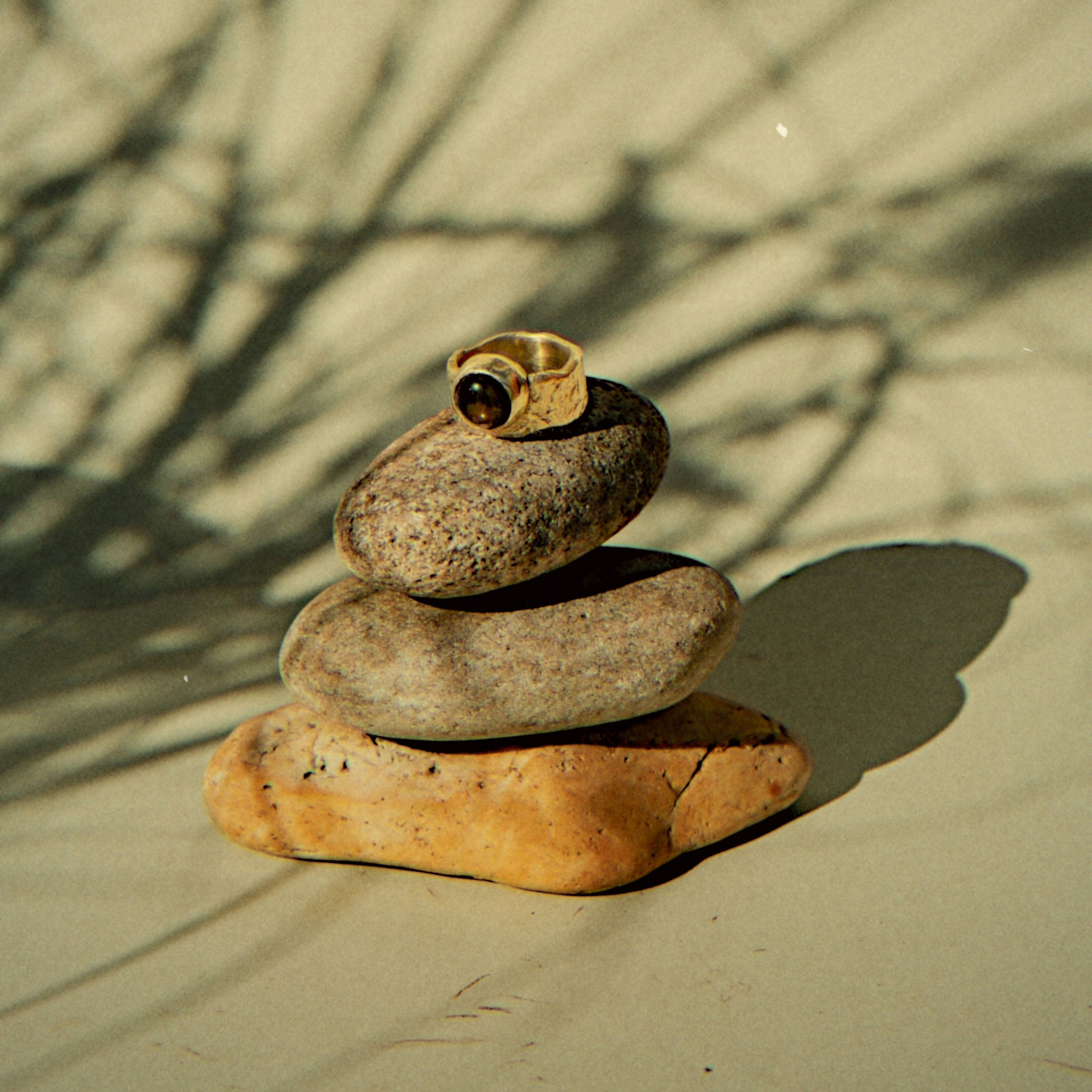 A gold ring with a dark stone is balanced on top of a stack of four smooth river stones on a beige surface, with leaf shadows cast across the background.