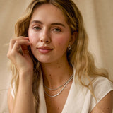 A blonde woman with freckles wearing a white draped top and layered silver necklaces, posing against a neutral beige background.