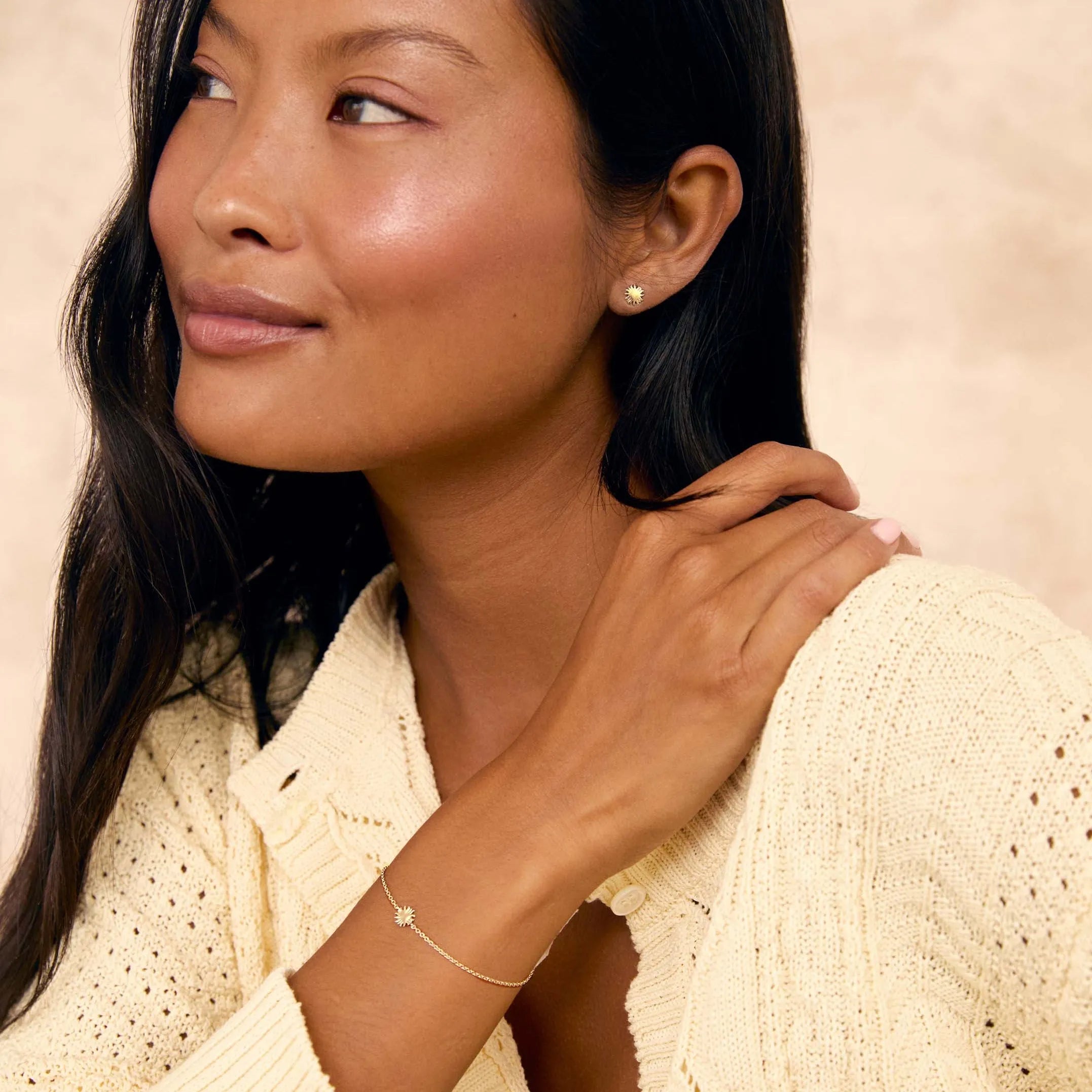 A woman with long dark hair wearing a cream-colored knit cardigan and a delicate gold sun-charm bracelet and matching studs, posing against a textured beige background.