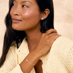 A woman with long dark hair wearing a cream-colored knit cardigan and a delicate silver sun-charm bracelet and matching studs, posing against a textured beige background.