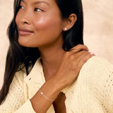 A woman with long dark hair wearing a cream-colored knit cardigan and a delicate silver sun-charm bracelet and matching studs, posing against a textured beige background.