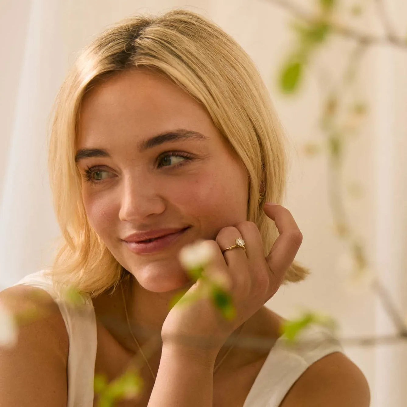 blond woman wearing a solitaire diamond engagement ring with blurred branches in for foreground