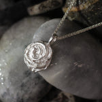 A silver pendant necklace with a textured, spiral rose design, resting on smooth gray river stones.