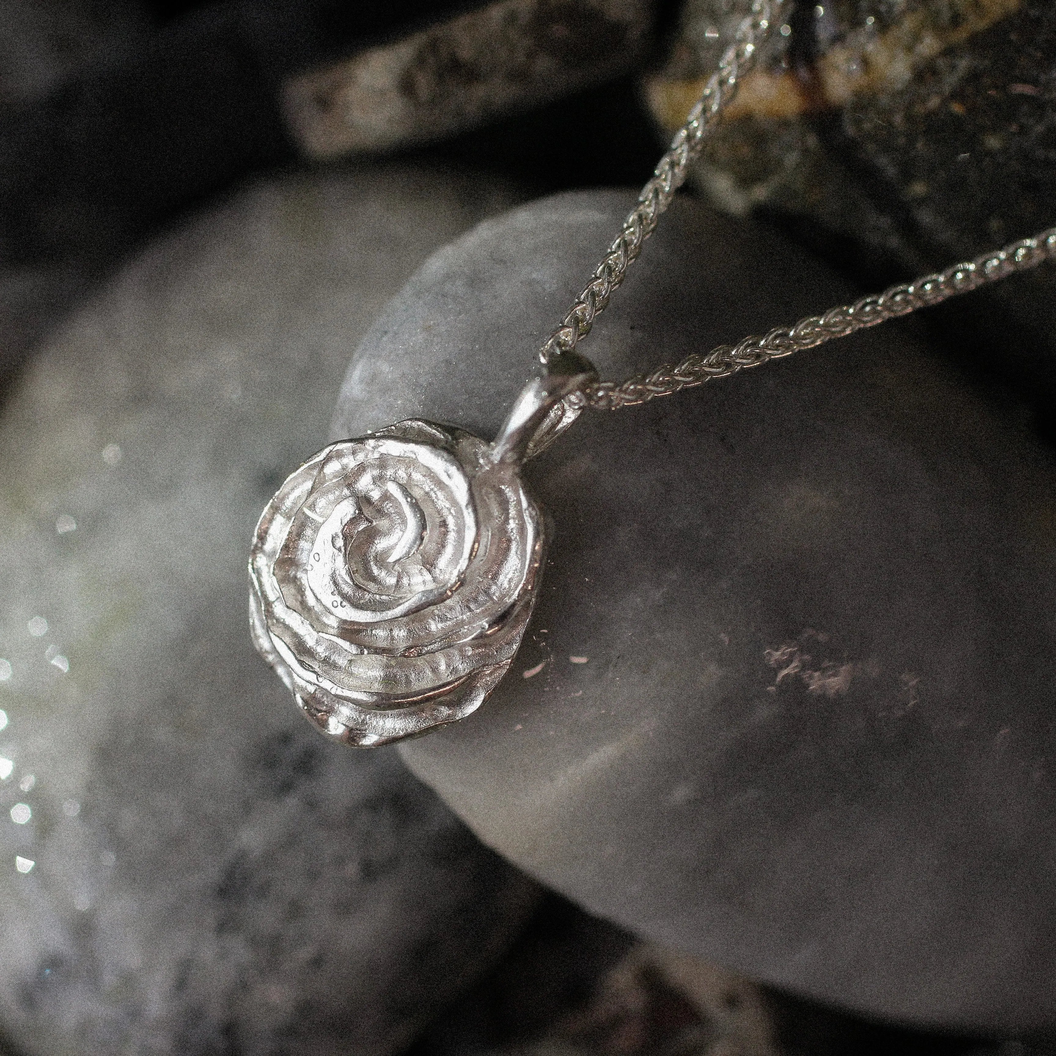 A silver pendant necklace with a textured, spiral rose design, resting on smooth gray river stones.