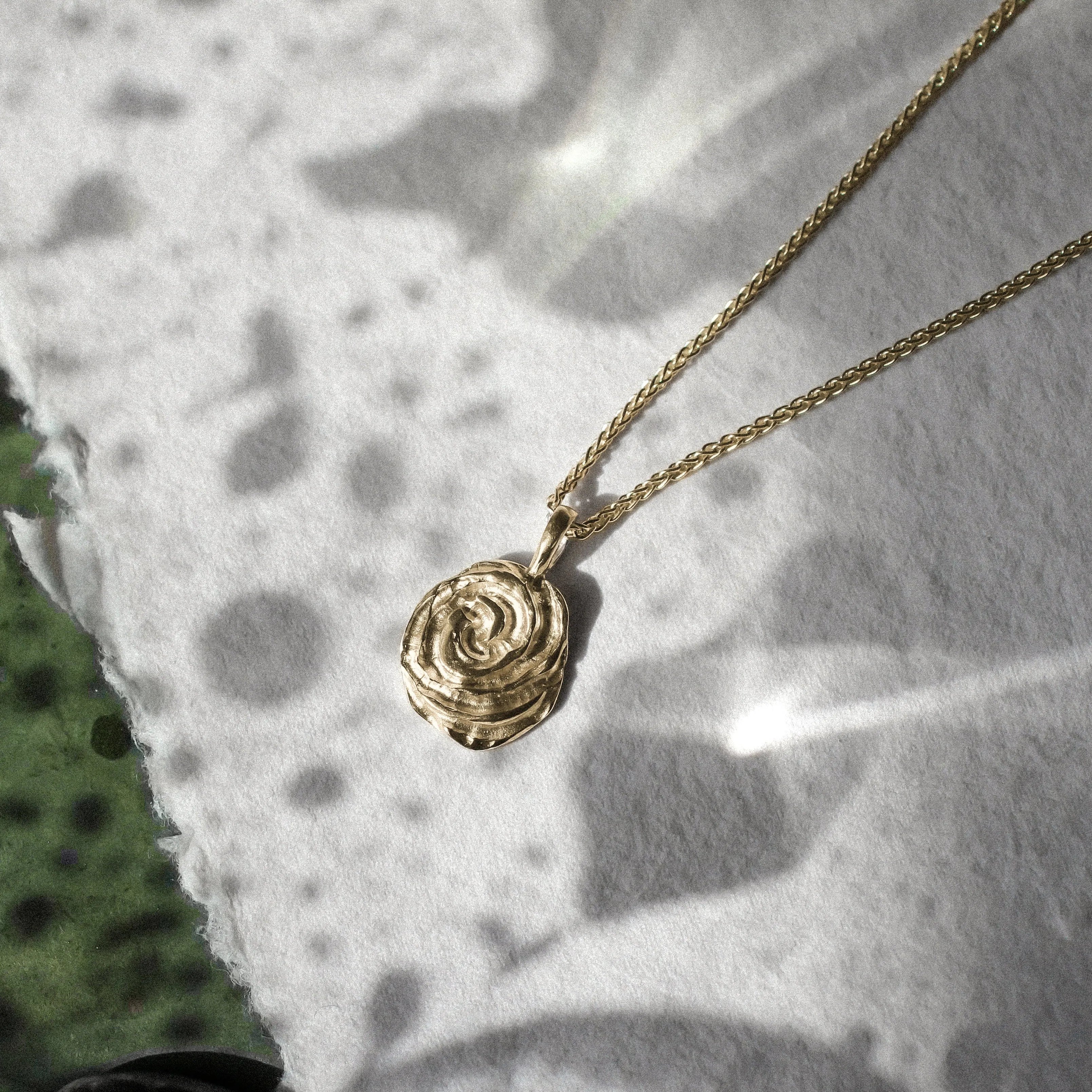 A delicate gold chain necklace with a small textured rose-shaped pendant, resting on torn white paper with a green mottled surface in the background.