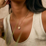 A close-up lifestyle shot of a woman wearing a delicate gold chain necklace with a textured circular medallion spiral pendant, paired with a light beige linen tank top.