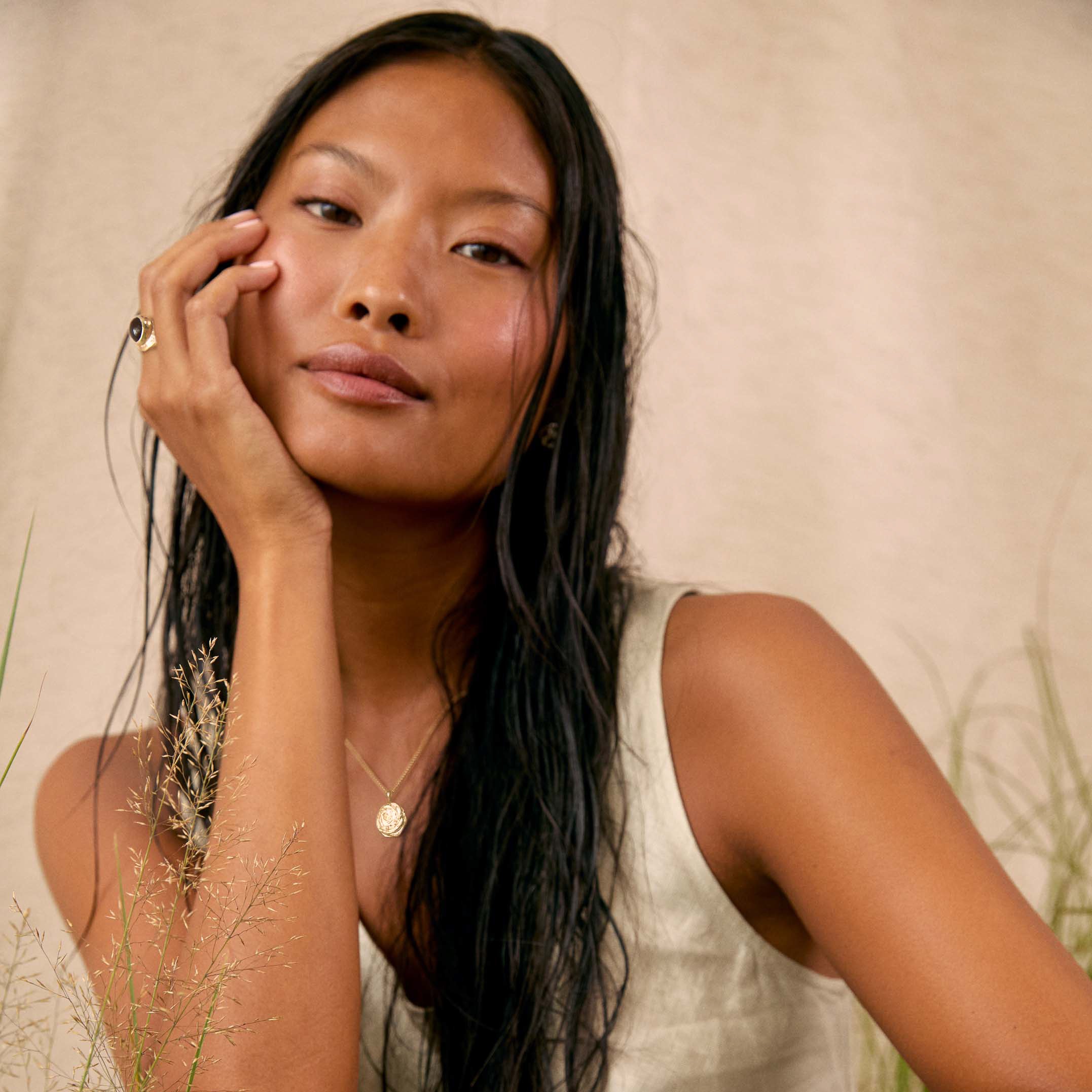 A woman with long dark hair resting her head on her hand, wearing a cream-colored sleeveless top, a delicate gold pendant necklace and chunky gemstone ring, set against a soft beige textured background with dried grass elements.
