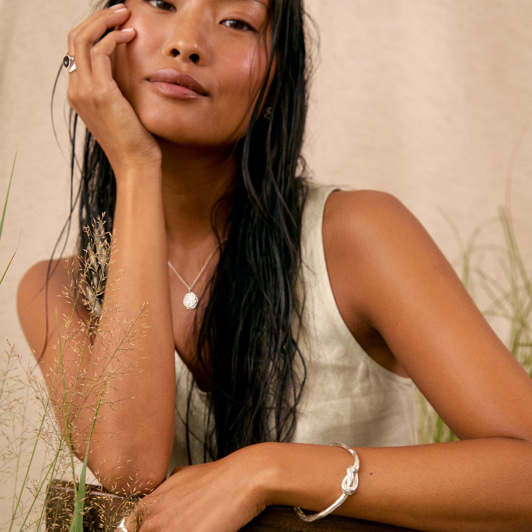 A woman with long dark hair rests her head on her hand, wearing a white sleeveless top and delicate silver jewelry including a necklace, bracelet, and rings, posed against a neutral beige background with dried grass.